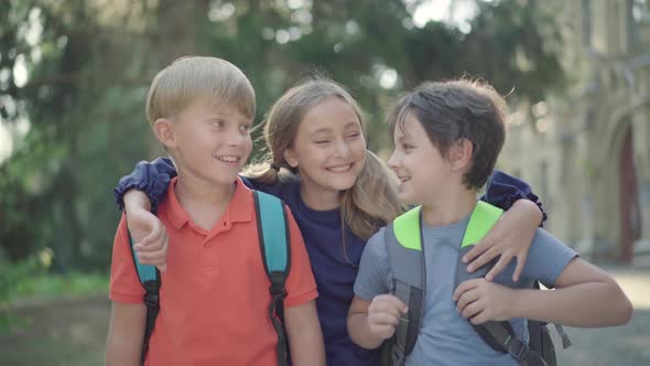 Cheerful Girl Running Along Sunny Schoolyard and Hugging Boys. Portrait of Three Joyful Caucasian