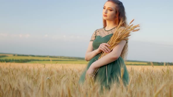 Beautiful woman in green dress on field with sheaf of wheat in hand at sunset light