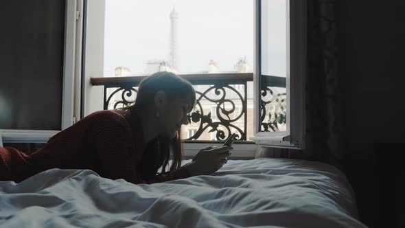 Inspiration Concept, Happy Blogger Woman Using Smartphone App Writing Post on Bed with Eiffel Tower