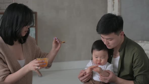 Happy father holds baby son on his lap while an Asian mom with spoon feeds baby