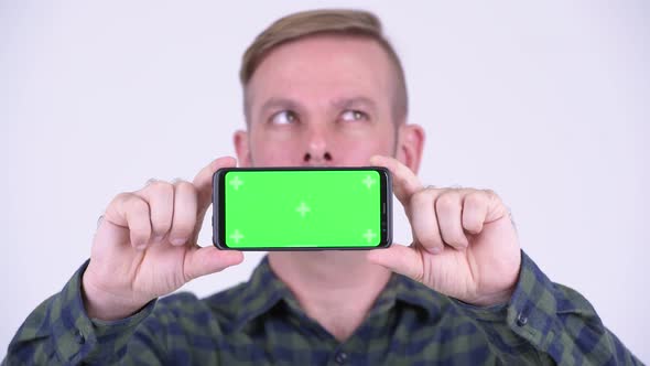 Portrait of Happy Blonde Hipster Man Thinking While Showing Phone