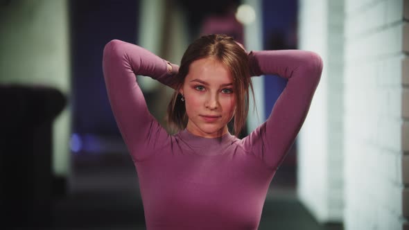 Young Pretty Woman in Purple Sports Suit Puts Her Hair in a Ponytail in the Gym Looks in the Camera