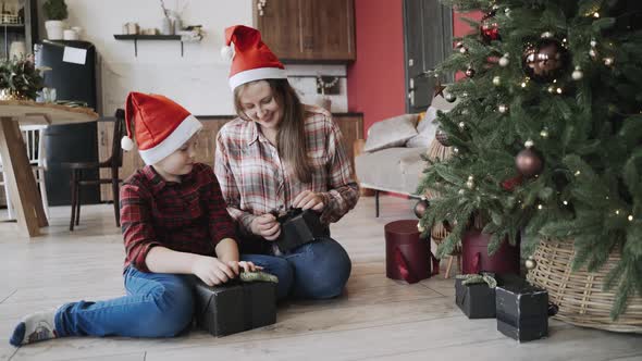 Happy Caucasian Mom and Son Sit Near Christmas Tree and Admire Gifts in Boxes