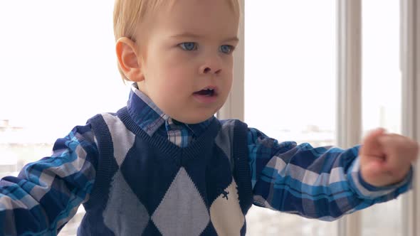 Cute Little Boy Points Finger And Takes In Mouth. Winter City View Through Windows