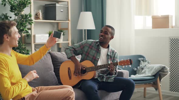 Two Young Men Have Fun Playing Guitar and Recording Video on Smarthphone at Home