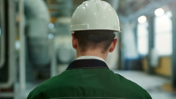 Backview of Engineer in Hardhat Walking in Factory and Writing Down on Clipboard