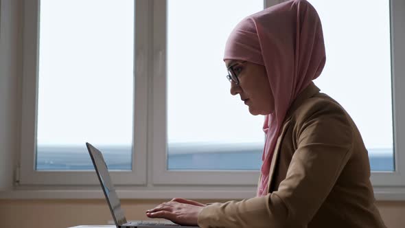Woman in Pink Hijab Typing on Laptop Against Window Background