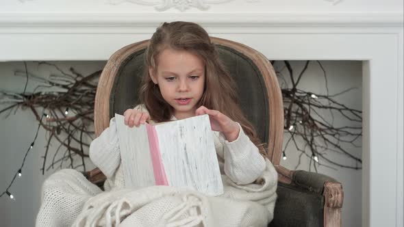 Lovely Little Girl Wrapped in White Christmas Blanket Reading a Book Sitting in an Armchair Near the