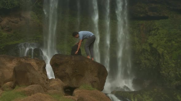 Guy with Backpack Stands on Rock Drinks Water By Waterfall