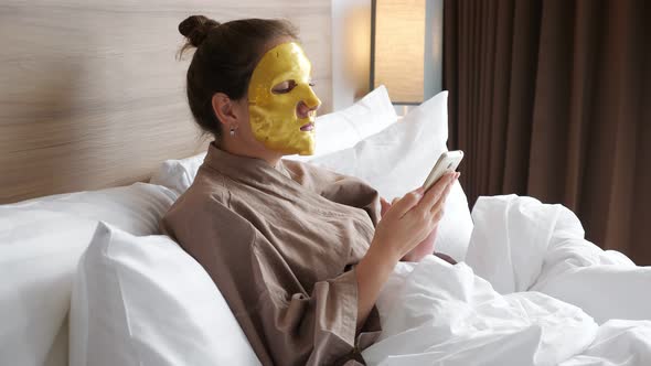 Girl with Golden Face Mask Uses Smartphone in Soft Bed