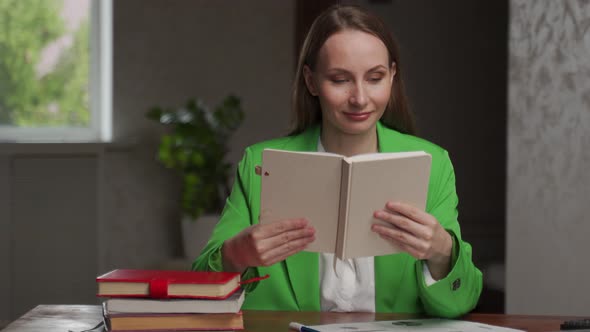 Smiling Young Woman Reads Interesting Book Sitting at Table