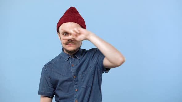 Portrait of Amusing Guy 20s Wearing Trendy Mustache and Hat Showing Peace Sign with Fingers While