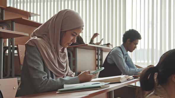 Female Muslim Student Sitting by Desk Using Smartphone