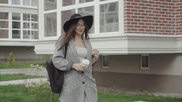 Pretty Woman in Black Hat and Glasses with Cup of Coffe Walking Down the Street