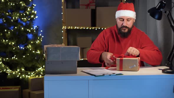 Dropshipping Owner Male Hindu Santa Claus Works at Night in His Home Office Knitting a Big Red Bow