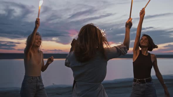 Group of Carefree Young Women Dancing on Beach with Fireworks at Sunset