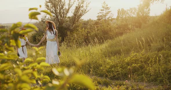 Two Girl Teenagers in White Dresses and Straw Hats with Flowers Bouquet Walking on Nature at Sunny