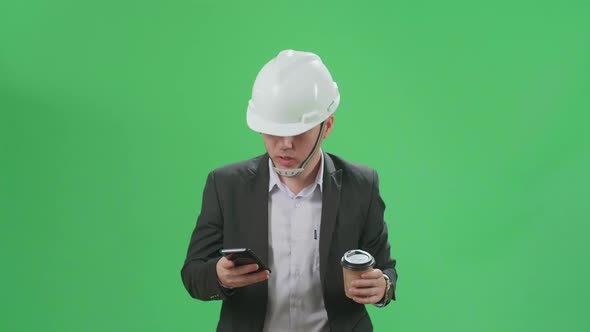 Asian Chief Engineer In The Hard Hat Use Mobile Phone And Drink Coffee While Walking On Green Screen