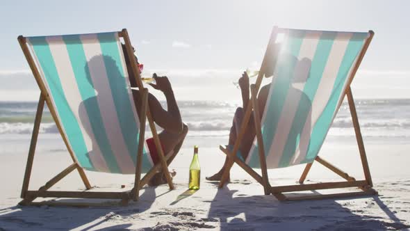 African american couple drinking wine together sitting on deck chairs at the beach