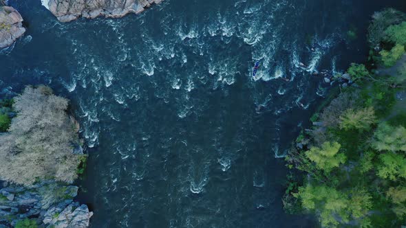 A Topdown Aerial View of a Kayak Floating on a Turbulent River Between Forest and Stone Cliffs