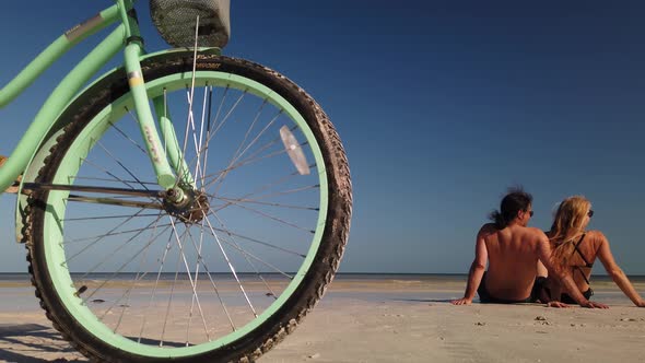 Wide shot of bike in foreground as man and woman lean back on their arms and talk while sitting in t