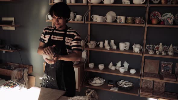 Young Male Potter Kneads Clay Against Background of a Rack with Ceramic Blanks