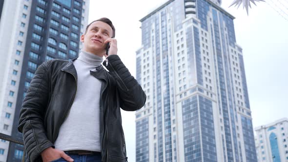 Man in a Leather Jacket Talking on the Phone Against the Backdrop of Tall Houses