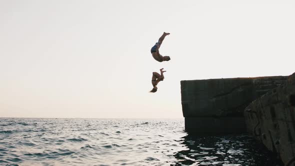 Young Woman and Man Synchronously Doing Frontflip From a Pier Into the Sea During Beautiful Sunrise