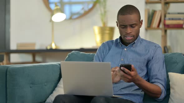 Young African Man Working on Smartphone and Laptop on Sofa 