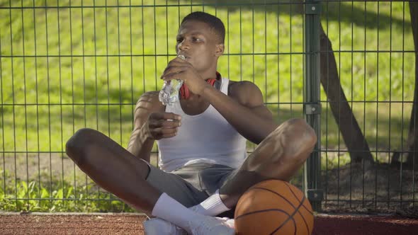 Portrait of Satisfied Basketball Player Sitting on Court with Ball and Drinking Fresh Cold Water