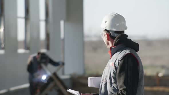 Male Engineer in Protective Helmet and in Safety Glasses with Drawing in Hand on Background 