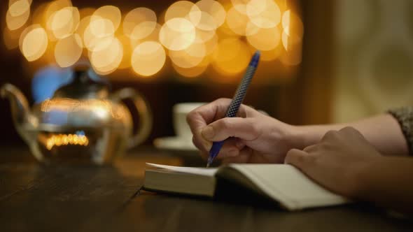 Woman Keeps a Diary While Sitting in Cozy Cafe with Cup of Tea. Female Hands Writes in Notebook