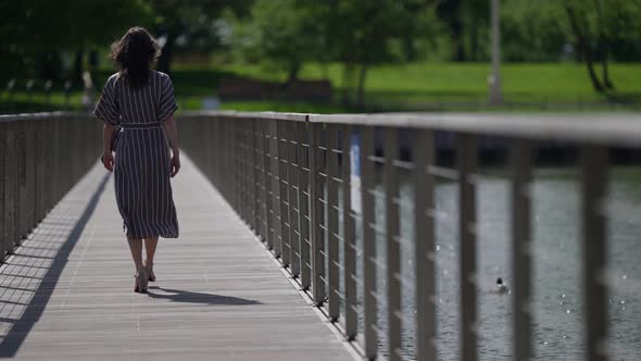 a Brunette in a Striped Dress and Highheeled Shoes Walks on a Pedestrian Bridge Over the River on a