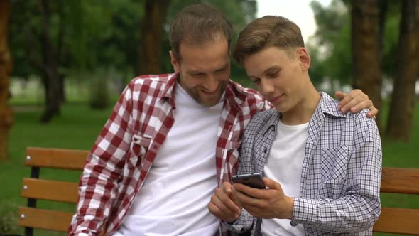 Male Teenager and Dad Watching Funny Video on Smartphone, Laughing and Smiling