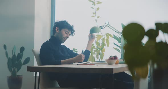 Young man sitting at table casually making notes while playing with plant
