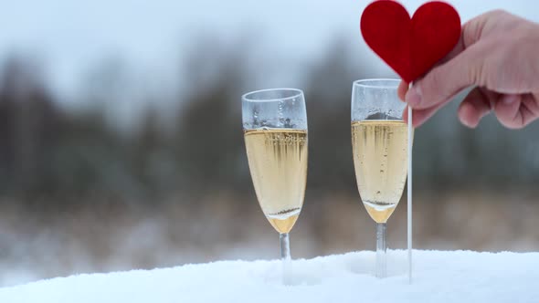 Mans Hand Put Heart on Stick Into Snow Near Glasses with Champagne