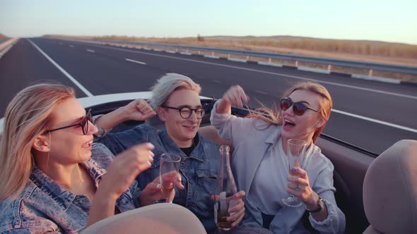Group of Friends Two Women and Man Sit in Back Seat of Convertible with Glasses and Bottle of