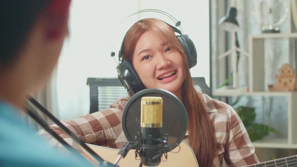 Young Asian Woman In Headphones Playing The Guitar And Singing While Recording Podcast With Male