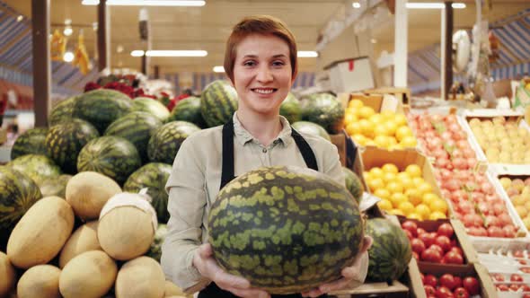 A vendor holds a watermelon and demonstrates fresh organic goods at a farmers market.