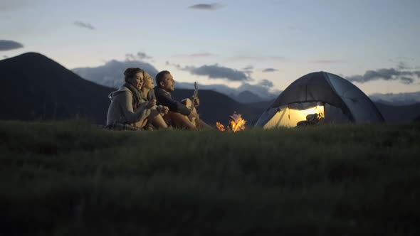 Group of Three Friends Play Guitar and Sing at Camp Fire in Nature Mountain Outdoor Camping Scene at