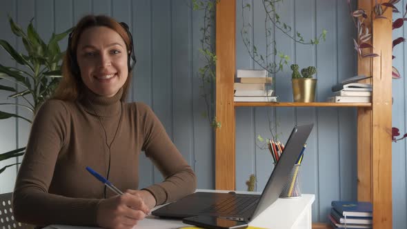 Woman Sit at Desk Look at Camera Hold Pen and Writes on Copybook Smile