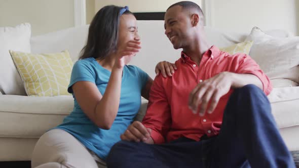 Happy biracial couple sitting on floor in living room, embracing and looking at camera