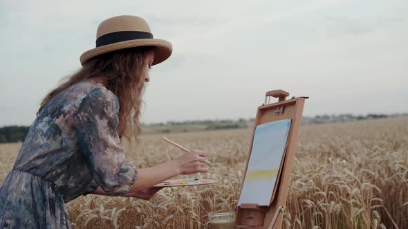 Pretty Paintress in Hat and Dress Painting Landscape on Canvas Among Wheat Field