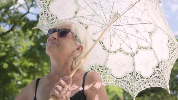 Portrait of Positive Smiling Mature Woman in Sunglasses Standing in the Park Under the White