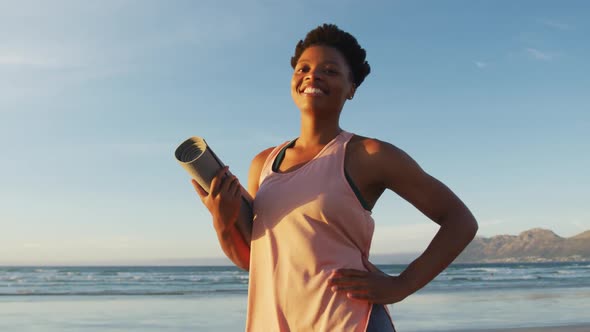 Portrait of african american woman holding yoga mat at the beach smiling