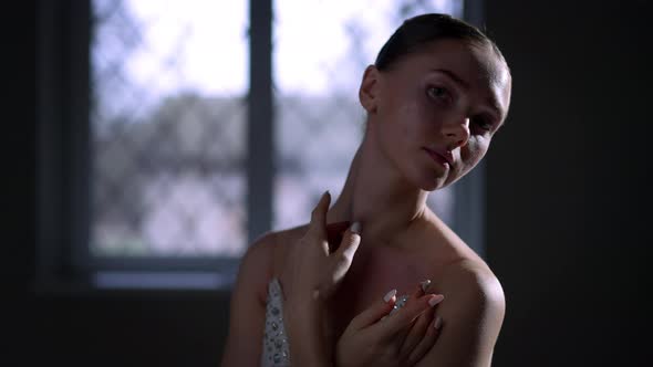 Charming Ballerina in White Dress Looking at Camera Standing in Darkness Indoors