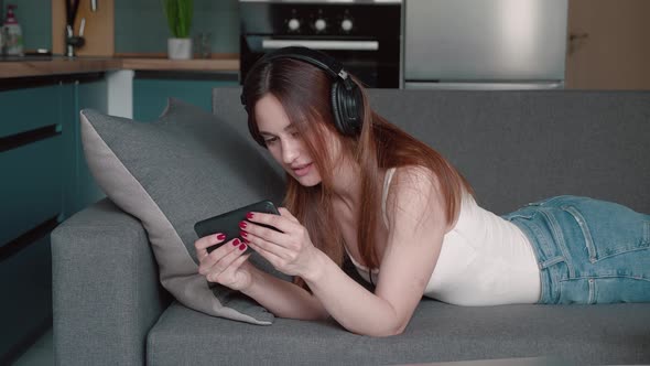 Funny Woman is Playing Video Game in Smartphone By Internet Using Headphones and Lying on the Couch