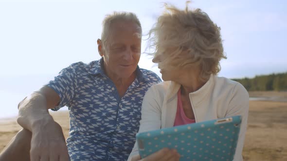 Happy Mature Couple Browsing Internet on Tablet Relaxing on Sea Beach