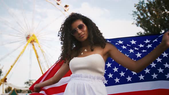 Afro American Woman in White Outfit and Sunglasses is Dancing Wrapping in Flag of USA Posing in Park