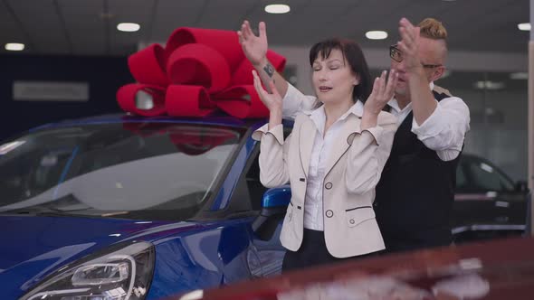 Loving Caucasian Husband Surprising Wife in Car Dealership Buying Blue Luxurious Vehicle with Large